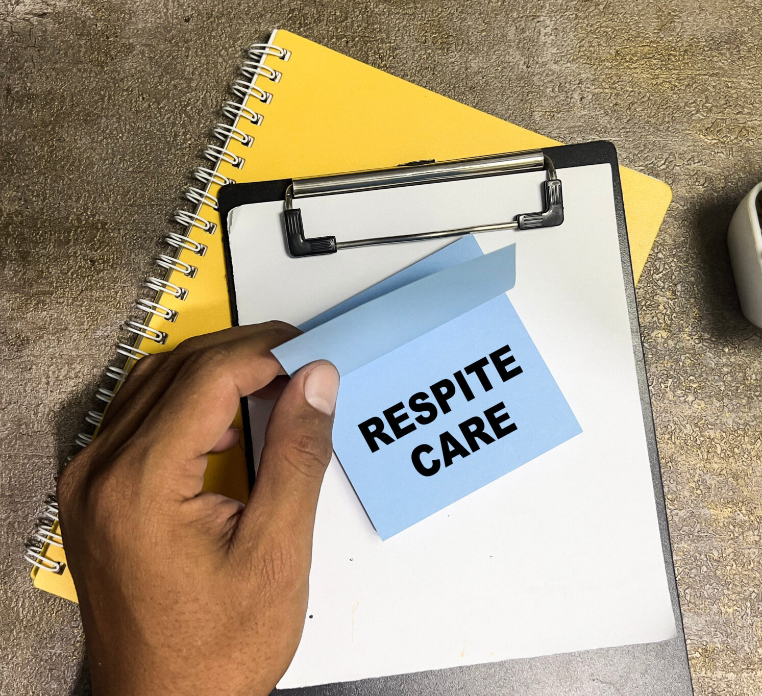 A hand holding a note labelled “Respite Care” above a clipboard and notebook, representing support services for caregivers and families.