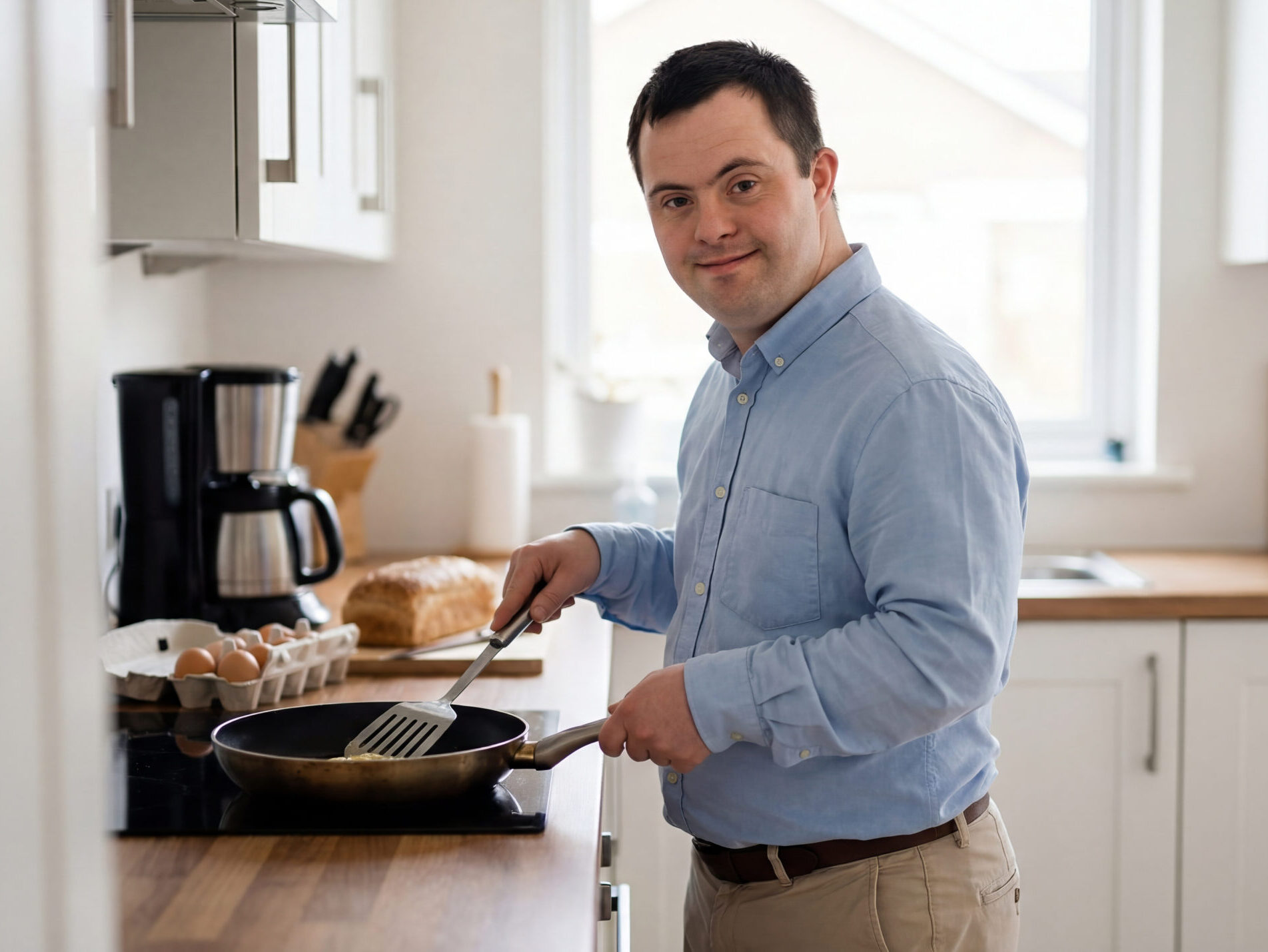 A man cooking at the stove in a bright kitchen, smiling while preparing food in a frying pan.