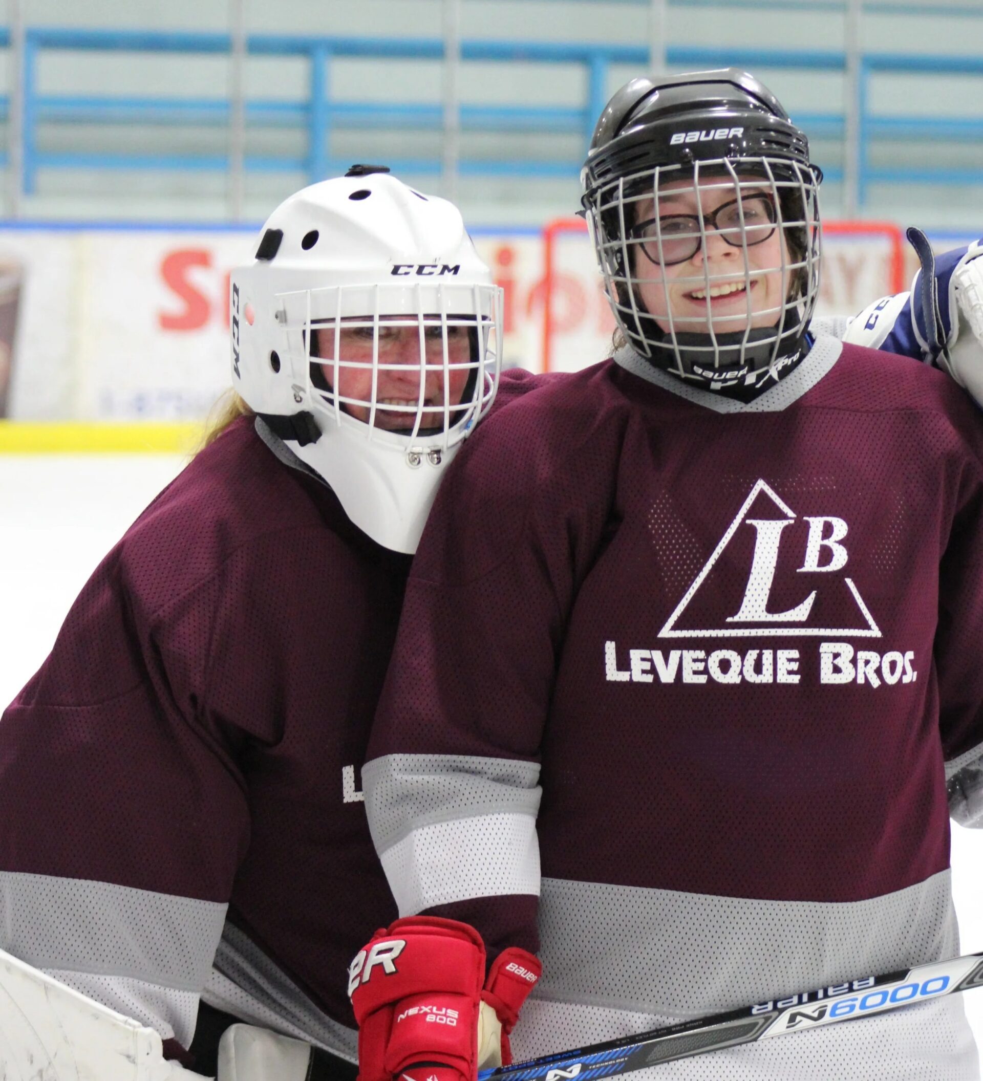 Two participants in hockey gear smiling together on an indoor ice rink during a community sports activity.