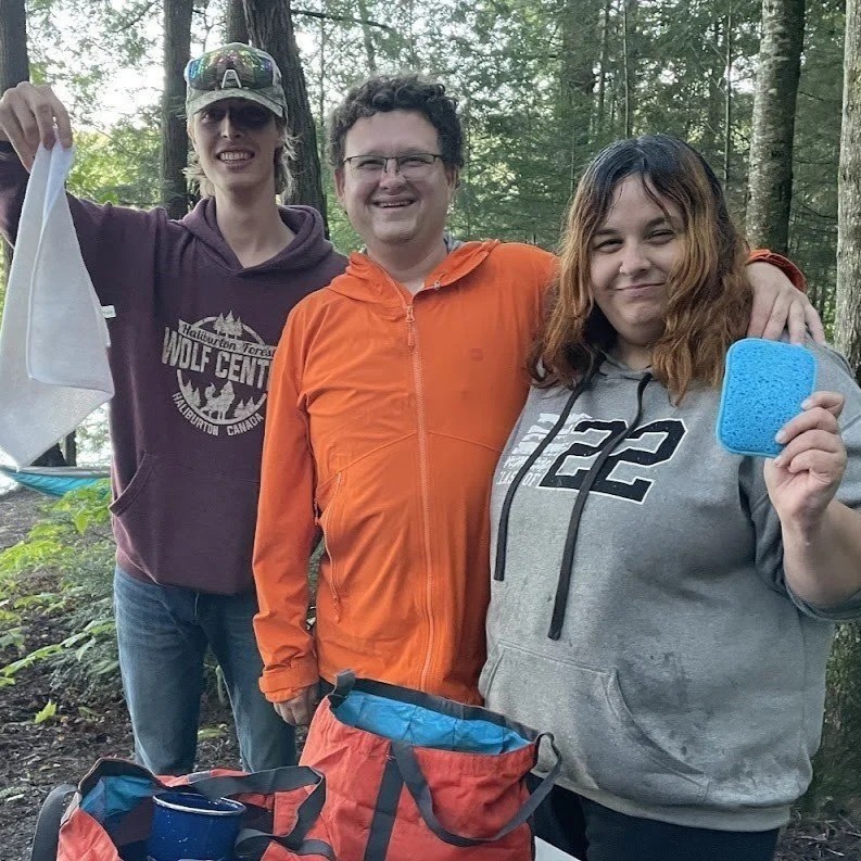 Three people smiling together at a lakeside campsite, holding cleaning supplies during an outdoor group activity.