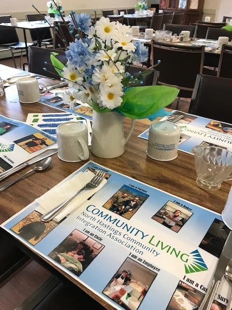 Table set for a community lunch with NHCIA Community Living materials and floral centrepieces.