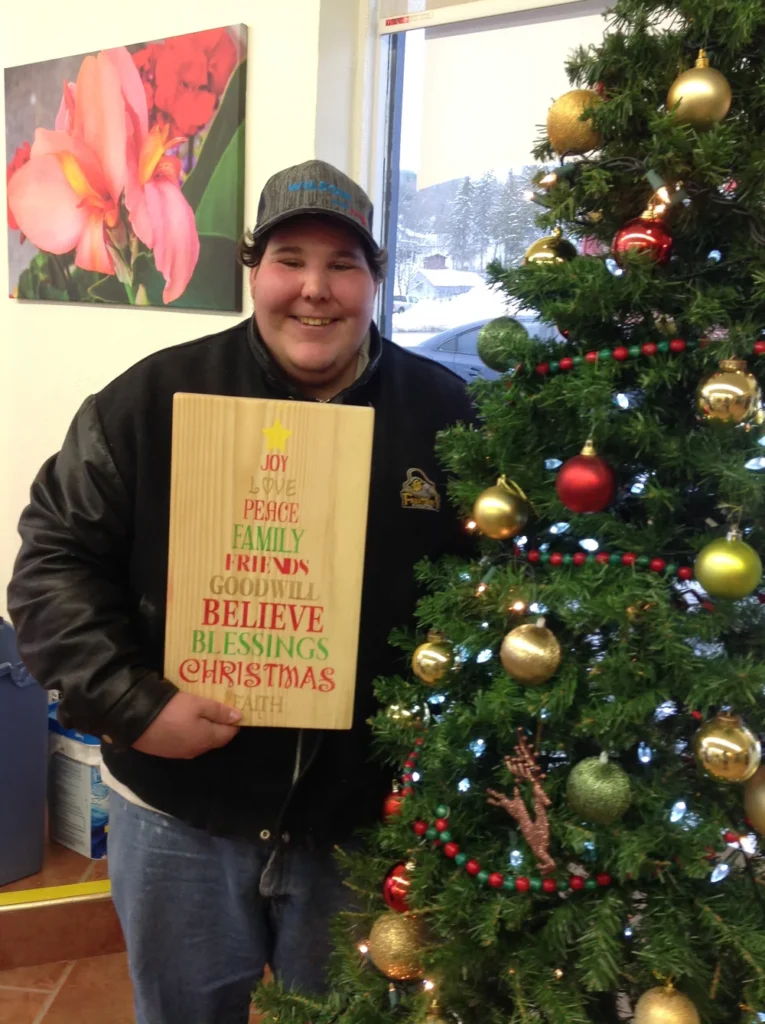 Participant holding a finished holiday sign beside a decorated Christmas tree at a Kairos Time event.