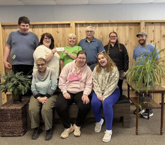Micheal and Pebbles from the Shamrock Club presenting a cheque to North Hastings Community Integration Association, surrounded by a group of smiling NHCIA members and staff indoors.